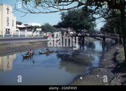 De jeunes enfants thaïlandais pagayent des canoës traditionnels le long d'un canal à Bangkok, en Thaïlande, en passant par une église chrétienne. 1968. Une passerelle en bois traverse le canal . Banque D'Images