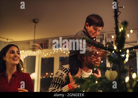 Garçon assis sur l'épaule de son père tout en décorant l'arbre de Noël. Famille décorant arbre de Noël ensemble à la maison. Banque D'Images