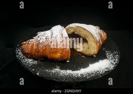 2 moitiés d'un croissant au chocolat, poudrées de sucre, sur une tuile de pierre noire, isolée sur fond noir. Banque D'Images