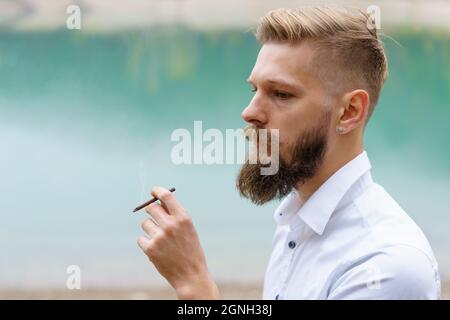 Beau gars sérieux, jeune homme sérieux fume, chauffage des produits du tabac, système à l'extérieur sur le fond du lac. Un garçon avec une cigarette dans la main dans le smog, la fumée, prend une bouffée. Mauvaise habitude. Banque D'Images