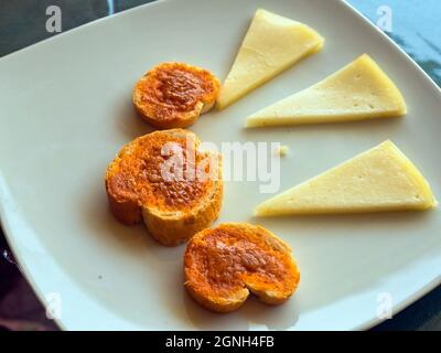 Vue en hauteur d'une plaque blanche avec trois tapas. Trois petits toasts ronds à la sauce canarienne rouge (mojo) et trois morceaux de fromage. Banque D'Images