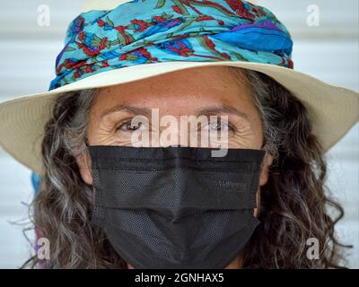 Charmante femme caucasienne positive mature avec de beaux yeux bruns porte chapeau panama avec élégant Blue Hat bande et noir masque jetable. Banque D'Images