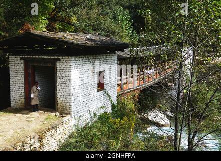 Un homme portant des vêtements traditionnels bhoutanais se dresse à l'entrée d'un ancien pont en porte-à-faux avec boiseries peintes colorées dans la campagne du Bhoutan Banque D'Images