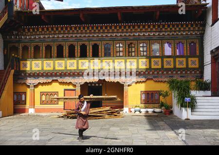 Un ouvrier portant un gho bhoutanais traditionnel porte du bois d'œuvre devant un bâtiment peint coloré sur le terrain de Tashichho Dzong à Thimphu, au Bhoutan. Banque D'Images