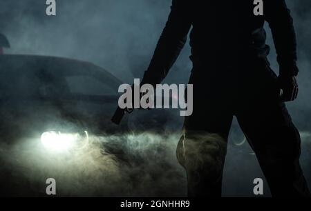 Un membre de gang avec une arme de poing devant sa voiture pendant les heures de nuit. Thème d'activité nuit illégal. Banque D'Images