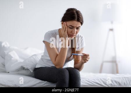 Déprimé triste triste malheureux jeune jolie femme européenne regardant le test négatif à la maison assis sur le lit dans la chambre à coucher à l'intérieur seul Banque D'Images