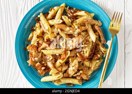 penne de pâtes alfredo de poulet à l'oignon caramélisé arrosé de pignons de pin et de parmesan râpé sur une assiette bleue sur une table en bois blanc, horizont Banque D'Images