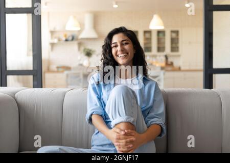 Portrait d'une jolie jeune femme brune souriant et regardant l'appareil photo, assise sur le couhc à la maison Banque D'Images