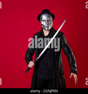 Un mystérieux assassin ninja dans un style noir. Un homme en vêtements noirs avec un cape et un chapeau, couvre son visage avec un masque et utilise un katana. Photo sur rouge Banque D'Images