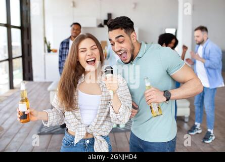 Jeune couple interracial chantant ensemble dans le microphone, tenant des bouteilles de bière, faisant la fête avec des amis à la maison Banque D'Images