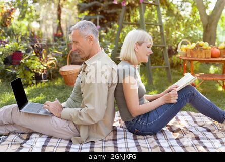 Les conjoints âgés se reposant dans le jardin, un homme utilisant un ordinateur portable, une femme lisant un livre, assis dos à dos, vue latérale Banque D'Images