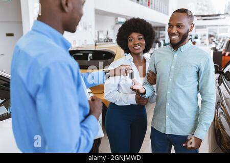 Vendeur donnant la clé de la voiture au jeune couple noir de la concession automobile. Concept de distribution locale du véhicule Banque D'Images