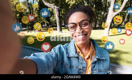 Jeune femme noire satisfaite en décontracté et lunettes fait de la photo en plein air pour les réseaux sociaux et le blog Banque D'Images