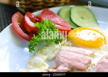 Œufs frits avec morceaux de bacon, tomates et concombres. Petit déjeuner sur une assiette blanche Banque D'Images