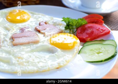 Œufs frits avec morceaux de bacon, tomates et concombres. Petit déjeuner sur une assiette blanche Banque D'Images