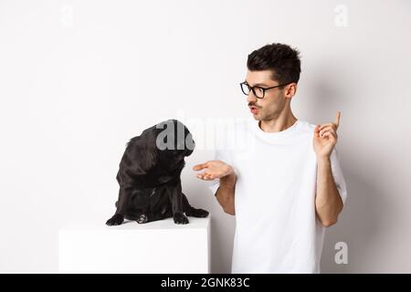 Beau jeune homme enseignant des commandes de chien, parlant à mignon pug noir, debout sur fond blanc Banque D'Images