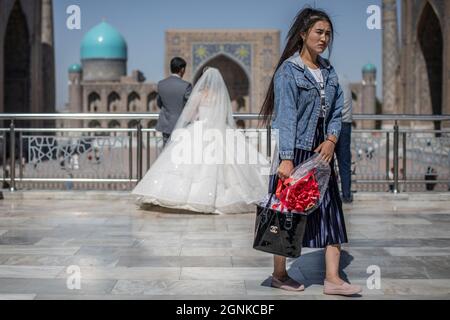 26 septembre 2021, Ouzbékistan, Samarkand : une femme ouzbek tient des fleurs alors que les jeunes mariés ont pris des photos au monument historique du Registan à Samarkand. Le Registan était une place publique et un centre de l'ancien Samarkand pendant l'Empire Timurid, utilisé pour des proclamations royales, annoncées par des explosions sur d'énormes tuyaux de cuivre et des exécutions publiques. Photo: Oliver Weiken/dpa Banque D'Images