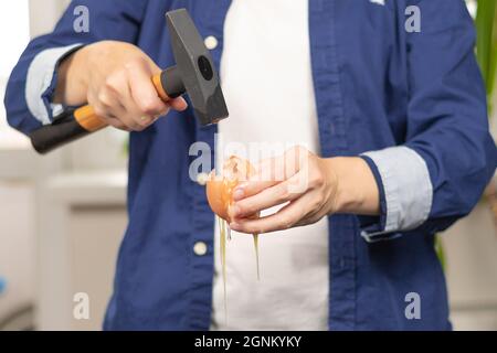 Les mains des femmes dans une chemise bleue cassent un œuf avec un marteau à la maison dans la cuisine. Mise au point sélective. Photo conceptuelle. Gros plan Banque D'Images