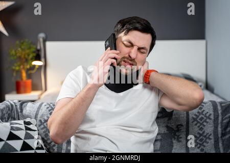 Handicapé homme douloureux avec un corset de cou parlant au téléphone à la maison. Homme avec un traumatisme de colonne vertébrale dans le cou étrésillon cervical appeler compagnie d'assurance pour l'aide médicale Banque D'Images