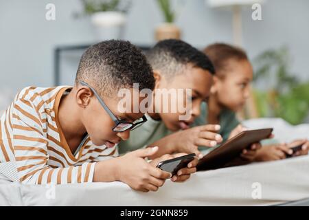 Vue latérale portrait de trois enfants dans la rangée à l'aide de gadgets tout en étant allongé sur le lit à la maison, espace de copie Banque D'Images