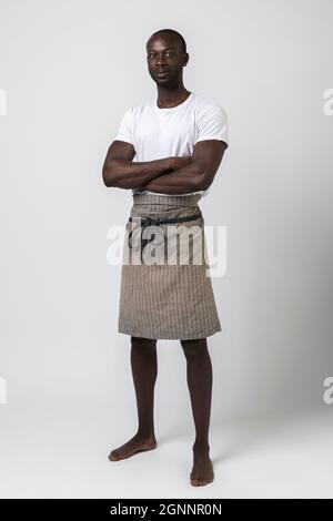 Un garçon africain avec un tablier pose trois quarts sur fond blanc en studio photo. Portrait du corps entier. Espace de copie, bras croisés Banque D'Images