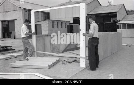 Années 1960, historique, deux hommes érigeant, à des fins de démonstration, un garage préfabriqué sur un terrain ouvert à l'extérieur d'un local industriel léger près de Witney, Oxford, Angleterre, Royaume-Uni, les travailleurs masculins tiennent le cadre de porte en bois, le fixant au niveau inférieur de panneaux en béton préfabriqué déjà boulonnés et fixés en position. Avec la croissance rapide de la propriété de voiture, il y a eu un deman pour loger des voitures de la météo et des bâtiments de garage domestique érigés à partir de panneaux de béton préfabriqués est devenu un site commun en Grande-Bretagne dans cette époque. Banque D'Images