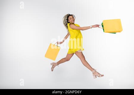 femme afro-américaine excitée en robe jaune lévitant avec des sacs de shopping isolés sur blanc Banque D'Images