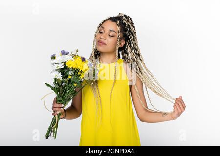 heureuse femme afro-américaine touchant des dreadlocks tout en regardant des fleurs colorées isolées sur blanc Banque D'Images