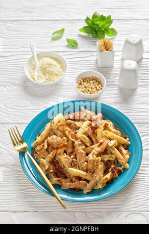 poulet alfredo pasta penne à l'oignon caramélisé arrosé de pignons de pin et de parmesan râpé sur une assiette bleue sur une table en bois blanc, à la verticale Banque D'Images