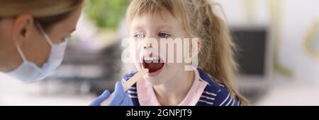 Otorhinolaryngologiste médecin en masque médical protecteur examinant la gorge de la petite fille avec une spatule en bois Banque D'Images