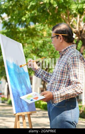 Inspiré asiatique mature homme dans une tenue décontractée debout sur le chevalet et travaillant sur la photo dans le parc Banque D'Images