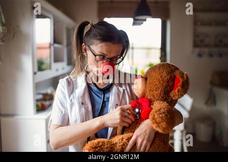 Portrait d'une femme drôle de médecin avec le nez de clown rouge tenant l'ours en peluche. Banque D'Images