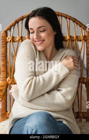 femme souriante dans un confortable chandail se embrassant dans un fauteuil en osier Banque D'Images