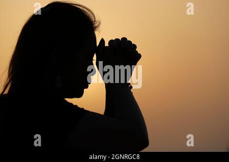 Silhouette au coucher du soleil de la femme fidèle priant, les mains se replient dans l'adoration à dieu. Concept de religion, de foi, de prière et de spiritualité. Banque D'Images