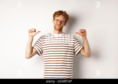 Un jeune homme plein d'assurance dans des lunettes aux cheveux rouges, pointant vers lui-même et regardant comme un professionnel, fond blanc Banque D'Images