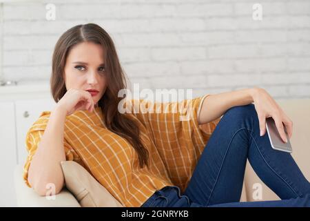 Jeune femme caucasienne pensive en blouse jaune et jeans assis sur un canapé dans la salle de séjour Banque D'Images