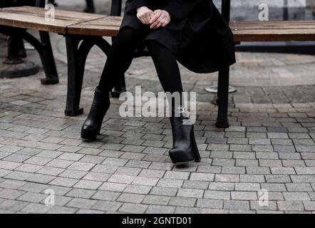 femme en manteau noir et bottes en cuir avec talon assis sur un banc Banque D'Images