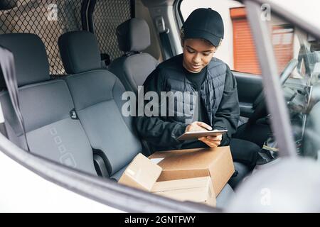 Femme dans un uniforme d'une compagnie de livraison vérifiant les informations sur une tablette numérique tout en étant assis sur le siège du conducteur Banque D'Images