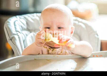 Bébé fille de 6 mois à la maison en chaise haute manger de la nourriture savoureuse à la maison. Bébé drôle avec le visage de purée de brocoli sain souillé tient la cuillère dans les mains Banque D'Images