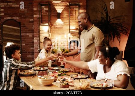 Portrait aux tons chaleureux de la famille afro-américaine en verre et en toaster tout en dégustant un dîner ensemble le soir Banque D'Images