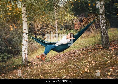 Jeune femme profitant de la chaleur de l'automne et se détendre dans le hamac balançant entre les arbres de bouleau. Loisirs en plein air hors-ville dans la nature concept im Banque D'Images