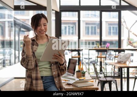 Une jeune femme asiatique se réjouit de la réussite ou de la bonne posture lorsqu'elle tient une tablette numérique à la main, une femme travaillant dans son bureau Banque D'Images