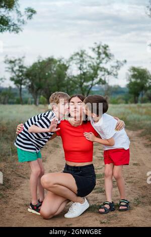 Portrait de famille authentique sur toute la longueur. Jeune maman décontractée avec deux fils embrassant et embrassant. Mère et enfant marchant dans la campagne. La vie quotidienne. Activités quotidiennes. Banque D'Images