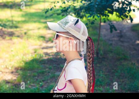Adorable petite fille avec de longs cheveux tressés en casquette de baseball en plein air sur un paysage ensoleillé de jour. Culture jeunesse style de vie photo authentique. Bon voyageur ch Banque D'Images
