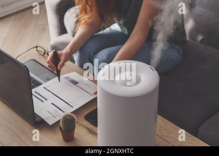 Femme indépendante utilise un humidificateur domestique sur le lieu de travail au bureau à domicile avec un ordinateur portable et des documents. Banque D'Images