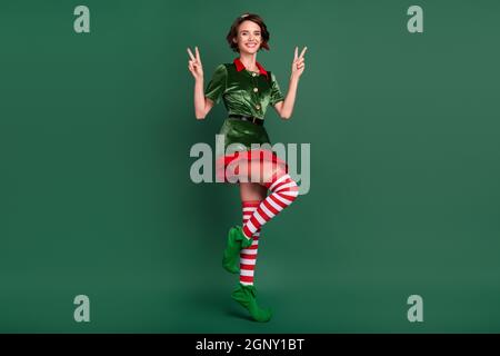 Photo de jolie charmante jeune femme habillée de costume d'elf montrant deux signes en V souriant isolé couleur vert fond Banque D'Images