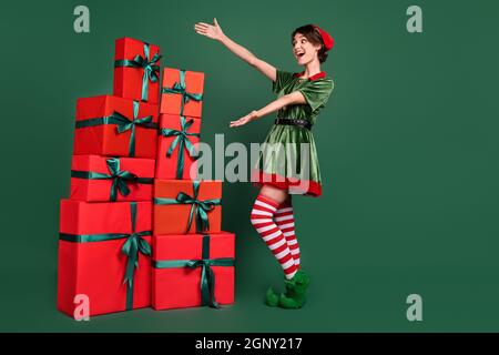 Photo de la jeune femme impressionnée confiante tenue d'elf montrant les bras boîtes cadeau pile sourire isolé couleur vert fond Banque D'Images