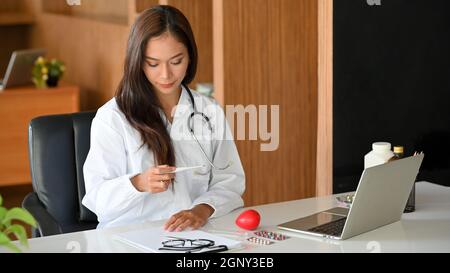 Femme médecin en robe blanche tenant ou regardant sur le thermomètre clinique pour enregistrer la température corporelle du patient Banque D'Images