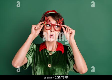 Photo d'une jeune femme assez choquée vêtue elf costume cerf lunettes regardant espace vide isolé couleur vert fond Banque D'Images