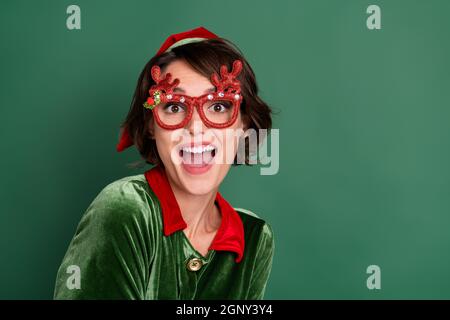 Photo de la jeune femme impressionnée vêtue elf costume cerf lunettes souriant espace vide isolé couleur vert fond Banque D'Images